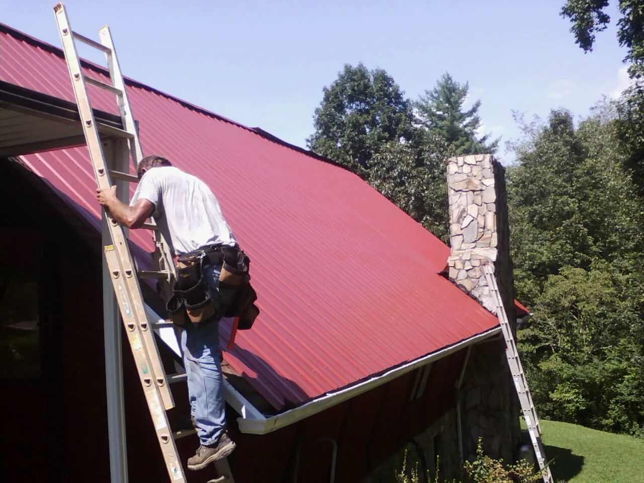 Roofer on a ladder beside a red metal roof and stone chimney — Soffit & Fascia Repair by Roofing Brothers in Elmwood Park